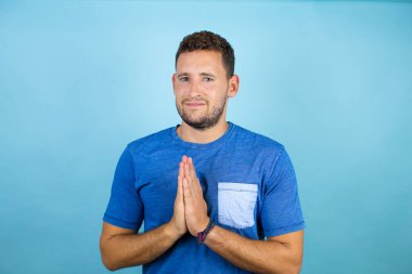 Mavi arka planda mavi tişört giyen yakışıklı genç adam elleri bağlı yalvarıyor ve dua ediyor. Yüzünde umut ifadesi var. Duygusal ve endişeli.