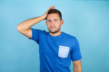 Mavi arka planda mavi tişört giyen genç yakışıklı bir adam bir elini kafasına koyup sanki bir şey unutmuş gibi gülümsüyordu.