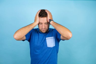 Mavi arka planda mavi tişört giyen yakışıklı genç adam yorgun ve elleri başının üstündeyken sıkılmış görünüyor.