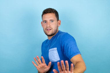 Mavi arka planda mavi tişört giyen genç yakışıklı adam korku ifadesinden korkuyor ve dehşete düşüyor. El hareketlerini durdur, şok içinde bağır. Panik konsepti.