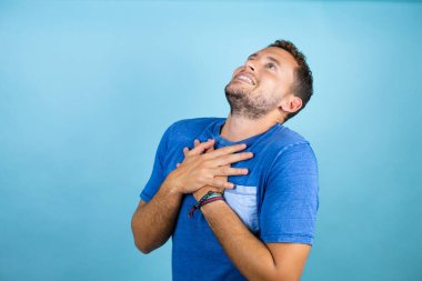 Mavi arka planda mavi tişört giyen genç yakışıklı adam elleri göğsünde ve yüzünde minnet dolu bir jest ile gülümsüyor..