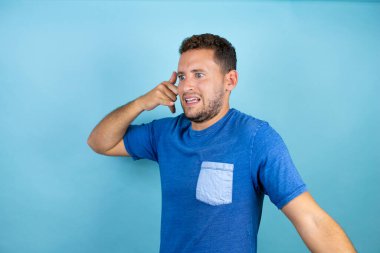 Mavi arka planda mavi tişört giyen genç yakışıklı adam telefon hareketlerini el ve parmaklarıyla yapıyor telefonda konuşuyor gibi.