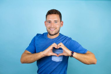 İzole edilmiş mavi arka planda mavi tişört giyen yakışıklı genç adam, ellerinde kalp sembolü ve şekli olan aşkla gülümsüyor. Romantik konsept.