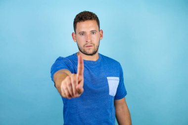 İzole edilmiş mavi arka plan üzerinde mavi tişört giyen genç ve yakışıklı bir numaralı parmaklarıyla işaret etmek ciddidir.