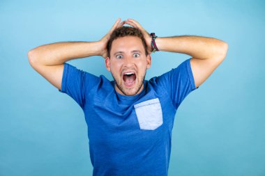 Mavi arka plan üzerinde mavi tişört giyen genç yakışıklı adam çılgın ve elleri kafasında korkmuş, şoktan korkmuş ve şaşırmış.