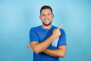 İzole edilmiş mavi arka plan üzerinde mavi tişört giyen genç yakışıklı adam gülümsüyor ve parmak tarafı olan bir tarafı işaret ediyor..