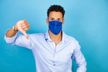 Soyutlanmış mavi arka planı olan mavi tişörtlü genç bir adam olumsuz bir ifade ve hoşnutsuzlukla başparmak hareketi yapıyor.