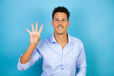 Mavi arka planda mavi tişört giyen genç yakışıklı dört numaralı parmaklarıyla işaret ederken kendinden emin ve mutlu gülümsüyor.