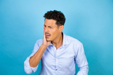 İzole edilmiş mavi arka planda mavi gömlekli genç adam diş ağrısı ya da diş ağrısı yüzünden acı dolu bir ifadeyle ağıza dokunuyor.