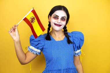 Sarı arka planda korkunç bir cadılar bayramı kostümü giyen kadın İspanyol bayrağını tutuyor.