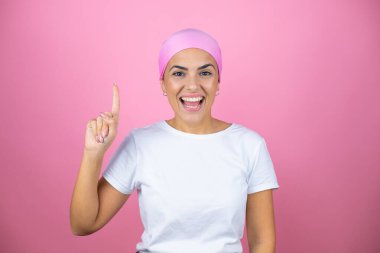 Pembe başörtüsü takan genç ve güzel bir kadın pembe arka planda kendini beğenmiş ve mutlu bir şekilde gülümserken bir numaralı parmaklarıyla işaret ediyor.