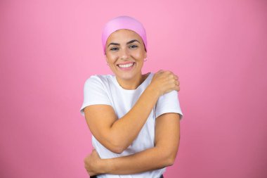 Pembe başörtüsü takan genç ve güzel bir kadın pembe arka planda kendine sarılıyor mutlu ve pozitif, kendinden emin gülümsüyor