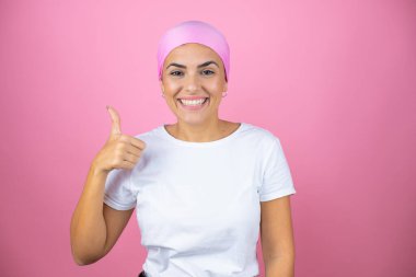 Pembe başörtüsü takan genç ve güzel bir kadın pembe arka planda gülümsüyor ve baş parmağıyla işaret veriyor.