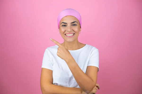 İzole edilmiş pembe arka planda pembe başörtüsü takan genç ve güzel bir kadın gülümsüyor ve elini ve parmağını kenarı işaret ediyor.