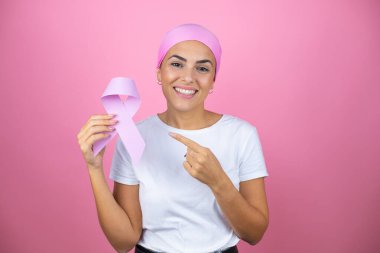 Genç ve güzel bir kadın pembe başörtüsü takıyor. Elinde kanser kurdelesi var. İzole edilmiş pembe arka planda.