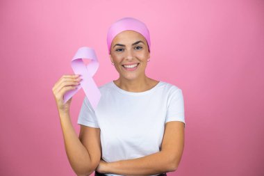 Genç ve güzel bir kadın pembe başörtüsü takıyor. Elinde kanser kurdelesi var. İzole edilmiş pembe bir arkaplan üzerinde.