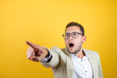 Genç yakışıklı iş adamı, izole edilmiş sarı arka planda takım elbise giyiyor. Parmağını işaret ediyor. İleriye şaşırmış, ağzı açık, şaşırmış bir ifade.