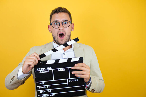 Young handsome businessman wearing suit over isolated yellow background surprised and holding clapperboard