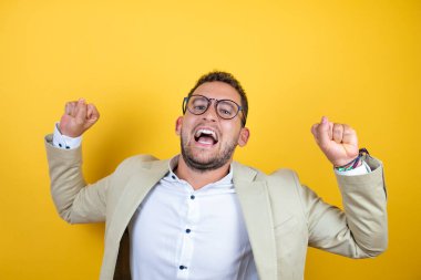 İzole edilmiş sarı arka planda takım elbise giyen yakışıklı genç işadamı mutlu ve heyecanlı bir şekilde kollarını kaldırıp, gülümseyerek ve başarı çığlıkları atarak kazanan bir jest yapıyor. Kutlama konsepti.