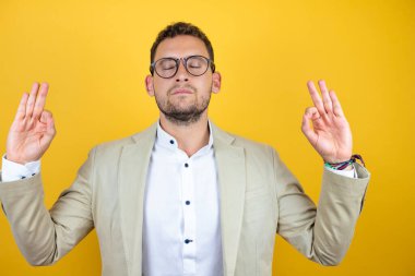İzole edilmiş sarı arka planda takım elbise giyen genç yakışıklı iş adamı rahatla ve parmaklarıyla meditasyon hareketi yaparken gözleri kapalı gülümse. Yoga konsepti.