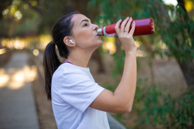 Ormanda spor yapan genç ve güzel bir kadın çok mutlu ve gülümseyen bir su içiyor.