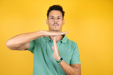 Sarı arka planda yeşil tişört giyen yakışıklı bir genç. Elleriyle, sinirli ve ciddi yüzüyle jest yapıyor.