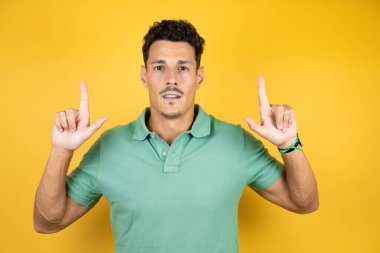 Sarı arka planda yeşil tişört giyen yakışıklı genç adam şaşırdı ve şaşırdı. Parmaklarıyla yukarı baktı ve kollarını kaldırdı.