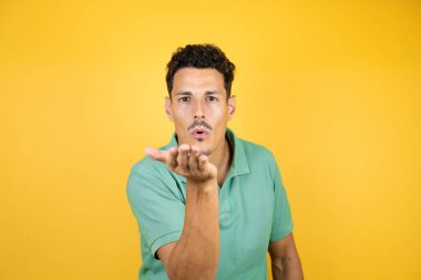 Sarı arka planda yeşil tişört giyen yakışıklı genç bir adam kameraya bakıp havada öpücükler savuruyor. Güzel ve seksi. Aşk ifadesi.