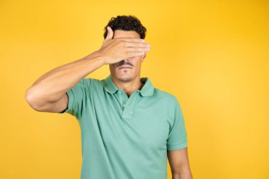 Sarı arka planda yeşil tişört giyen yakışıklı genç adam ciddi ve eliyle gözlerini kapatıyor.