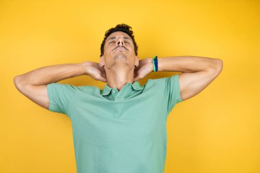 Yeşil tişört giyen yakışıklı genç adam izole edilmiş sarı arka planda rahatlıyor ve esniyor, kolları ve elleri başının arkasında ve boynunun arkasında mutlu bir şekilde gülümsüyor.