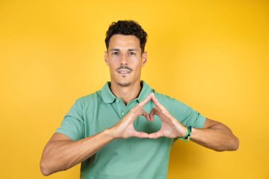 İzole edilmiş sarı arka planda yeşil tişört giyen yakışıklı genç adam kalp sembolünü ve şekillerini elleriyle göstererek gülümsüyor. Romantik konsept.