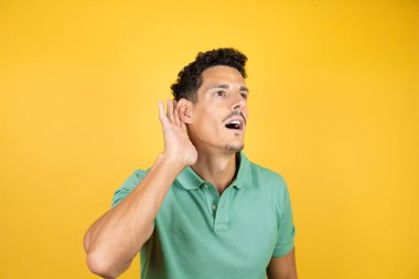 İzole edilmiş sarı arka planda yeşil tişört giyen yakışıklı bir adam kulağından girip kulağına fısıldayarak dedikodu ve dedikoduları dinliyor.