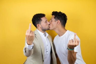 Young gay couple of two men wearing casual clothes and mask over isolated yellow background kissing and showing middle finger doing fuck you bad expression, provocation and rude attitude. screaming excited