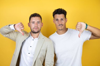 İzole edilmiş sarı arka planda, kızgın yüzlü, negatif işaretli, başparmağı aşağıda olan, gündelik kıyafetler giyen iki genç eşcinsel erkek.