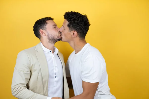 İzole edilmiş sarı arka planda sıradan kıyafetler giyen iki genç gay öpüşürken gülümseyerek duruyorlar.