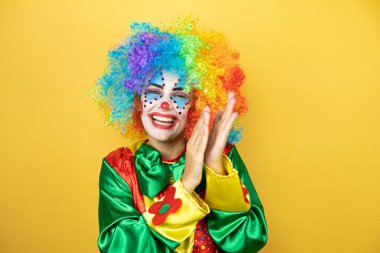 Sarı arkaplanda duran palyaço alkışlıyor ve alkışlıyor mutlu ve neşeli, gülümseyen gururlu eller birlikte.