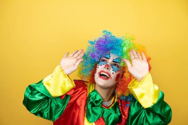 Sarı arkaplanda dikilen palyaço şok içinde yüzünü ve gözlerini kapayarak utangaç ifadesiyle parmaklarının arasından bakıyor.