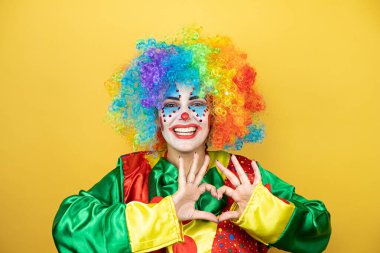 Sarı arkaplanda dikilen palyaço, sevgiyle gülümsüyor. Kalp sembolünü ve şeklini elleriyle gösteriyor.