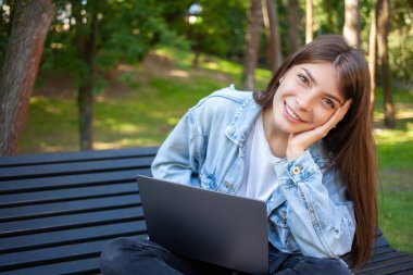 Teknoloji, eğitim ve insanların yaşam tarzı. Parktaki bankta dizüstü bilgisayarla oturan, gülümseyen ve kameraya bakan mutlu esmer genç kız. Yaz sabahı.
