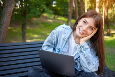 Halk yaşam tarzı, eğitim ve teknoloji kavramı. Serbest çalışan mutlu esmer kadın ya da öğrenci, parktaki bankta dizüstü bilgisayarla oturuyor, gülümsüyor ve kameraya bakıyor. Kopyalama alanı oluştur.