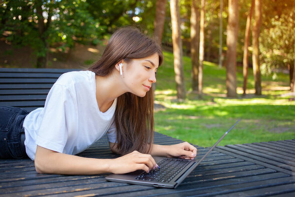 Remote working, education and technology concept. Young happy girl freelancer in wireless earphones, wearing white t-shirt, working and taking notes at laptop outdoors. Empty copy space.