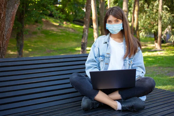 Quarantine, remote work and pandemic concept. A young European student female wearing medical mask for protection from virus, using laptop, sitting on the bench in park and looking to the camera. Empty copy space.