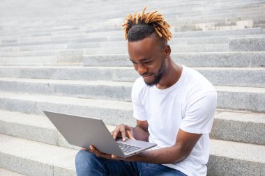 Serbest meslek, teknoloji ve eğitim kavramı. Dışarıda dizüstü bilgisayarda çalışan, parktaki merdivenlerde oturan konsantre olmuş genç bir Afro-Amerikalı. Boş kopyalama alanı.