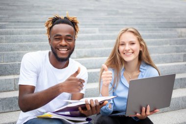 Serbest meslek ve eğitim kavramı, Afrikalı erkek ve Avrupalı kadın. Şanslı yüzlü öğrenciler, parktaki merdivenlerde oturuyor, ders projesi yapıyor ve kameraya bakıyor..