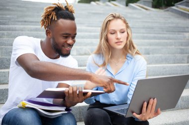 Eğitim, öğrenme ve insan konsepti. Genç, Afro-Amerikalı bir adam ve elinde dizüstü bilgisayarı olan Avrupalı bir kız. Parkta merdivenlerde oturuyor, sınavlarına çalışıyor ve bir ders projesi yapıyor..