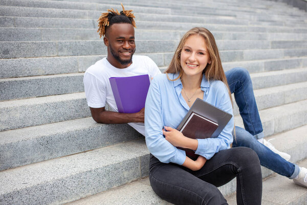 Portrait of two mixed race friends students with happy faces African American guy and European female sitting on stairs in the city park, rest after class, looking at a camera. Empty copy space.