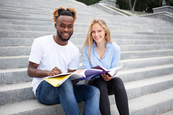 Education, learning and people concept - young, African American guy and European female sitting on stairs in the city park with smiling or happy faces, study for their exams and look to the camera.