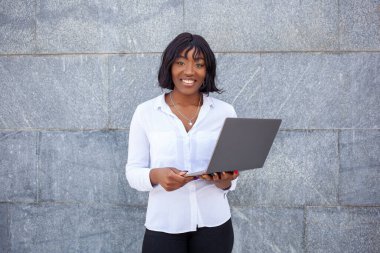 Mutlu yüzlü Afro-Amerikalı bir kadın bilgisayarını dışarıda çalışmak için kullanıyor, gri arka planda poz veriyor. Eğitim ve uzaktan çalışma kavramı.