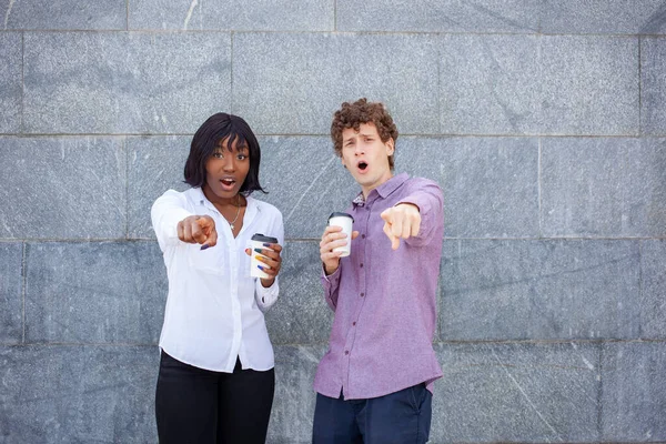 Multiracial friends hang out together, pose against gray background, looking and pointing fingers at camera. African American woman and European man are with surprise faces holding cup of coffee.