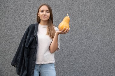 Gri arka plandaki kameraya bakarak, sol elinde olgun balkabağı olan, gündelik giysiler giyen genç, güzel Avrupalı bir kadın. Cadılar Bayramı partisi ve insanların konsepti.